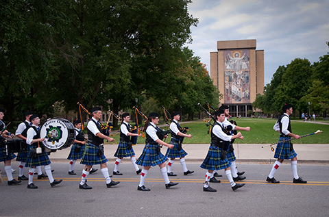 A pipe band in blue and green kilts marches on a road. Members play bagpipes and a large white drum with "Notre Dame" and a shamrock. Behind them is the Hesburgh Library, featuring the "Word of Life" mosaic.